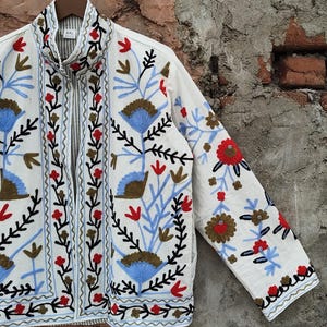 K&ouml;nnte beinhalten: Wei&szlig;e Jacke mit aufwendiger Blumenstickerei in Rot, Blau und Braun. Die Jacke hat einen hohen Kragen und einen durchgehenden Rei&szlig;verschluss. Die &Auml;rmel und die Vorderteile sind mit detaillierten gestickten Blumen und Bl&auml;ttern verziert.