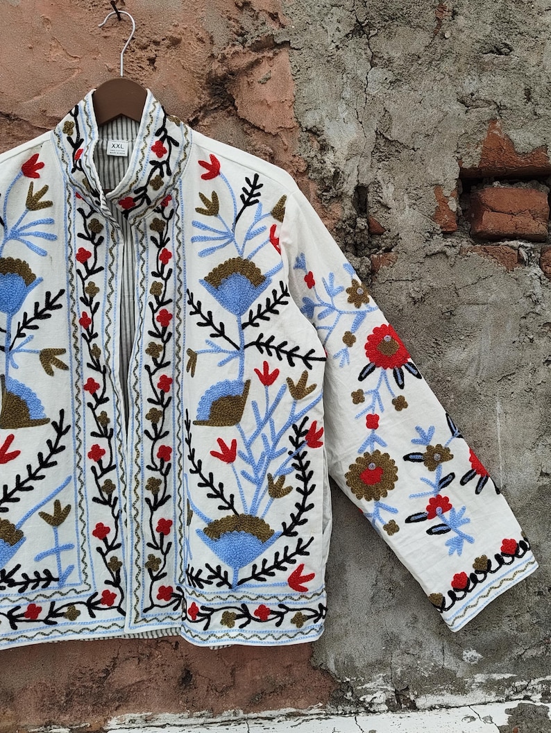 K&ouml;nnte beinhalten: Wei&szlig;e Jacke mit aufwendiger Blumenstickerei in Rot, Blau und Braun. Die Jacke hat einen hohen Kragen und einen durchgehenden Rei&szlig;verschluss. Die &Auml;rmel und die Vorderteile sind mit detaillierten gestickten Blumen und Bl&auml;ttern verziert.