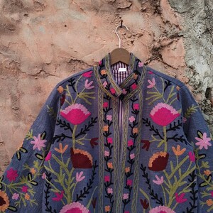 May include: A blue jacket with intricate floral embroidery in pink, orange, and green. The jacket features a high collar, a full zipper, and a striped lining. The floral design is detailed and covers the front and sleeves.