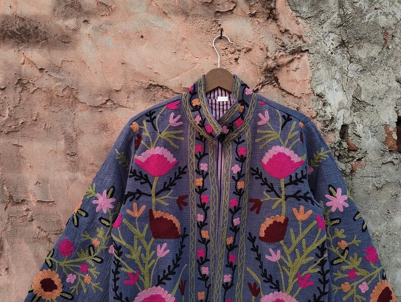 May include: A blue jacket with intricate floral embroidery in pink, orange, and green. The jacket features a high collar, a full zipper, and a striped lining. The floral design is detailed and covers the front and sleeves.