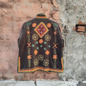 May include: A dark brown embroidered jacket with a floral design. The jacket features orange, green, and white floral patterns, with a diamond-shaped design on the back. The jacket is hanging on a green hanger.