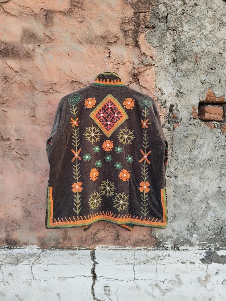 May include: A dark brown embroidered jacket with a floral design. The jacket features orange, green, and white floral patterns, with a diamond-shaped design on the back. The jacket is hanging on a green hanger.