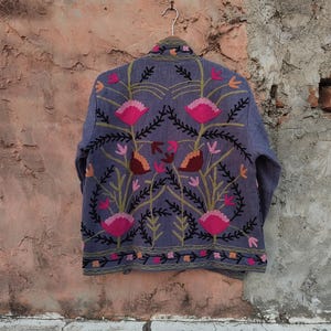 May include: A blue jacket with intricate floral embroidery in pink, orange, and green. The jacket is hanging on a metal hanger against a textured, neutral-toned wall. The embroidery features stylized flowers and leaves.