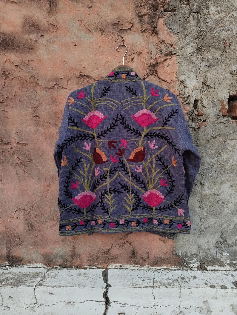 May include: A blue jacket with intricate floral embroidery in pink, orange, and green. The jacket is hanging on a metal hanger against a textured, neutral-toned wall. The embroidery features stylized flowers and leaves.