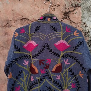 May include: A blue jacket with floral embroidery. The jacket features a detailed design of pink, orange, and brown flowers with green stems and black accents. The jacket is hanging on a metal hanger against a textured, brown wall.