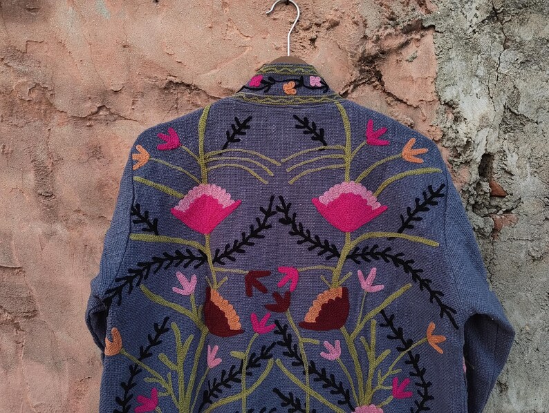 May include: A blue jacket with floral embroidery. The jacket features a detailed design of pink, orange, and brown flowers with green stems and black accents. The jacket is hanging on a metal hanger against a textured, brown wall.