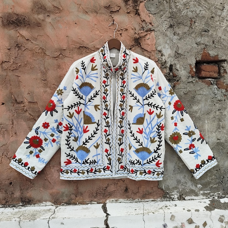 K&ouml;nnte beinhalten: Weisse Jacke mit farbenfroher Blumenstickerei. Die Jacke hat einen hohen Kragen und einen durchgehenden Rei&szlig;verschluss. Die Stickerei umfasst rote, blaue und braune Blumen und Bl&auml;tter. Die Jacke h&auml;ngt an einem Holzkleiderb&uuml;gel.