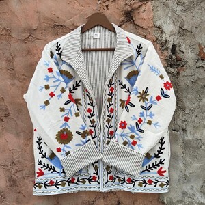 K&ouml;nnte beinhalten: Wei&szlig;e Jacke mit floralen Stickereien in Rot, Blau und Braun. Die Jacke hat ein gestreiftes Futter und Manschetten und h&auml;ngt an einem Holzb&uuml;gel. Die Stickerei zeigt eine Vielzahl von Blumen und Bl&auml;ttern.