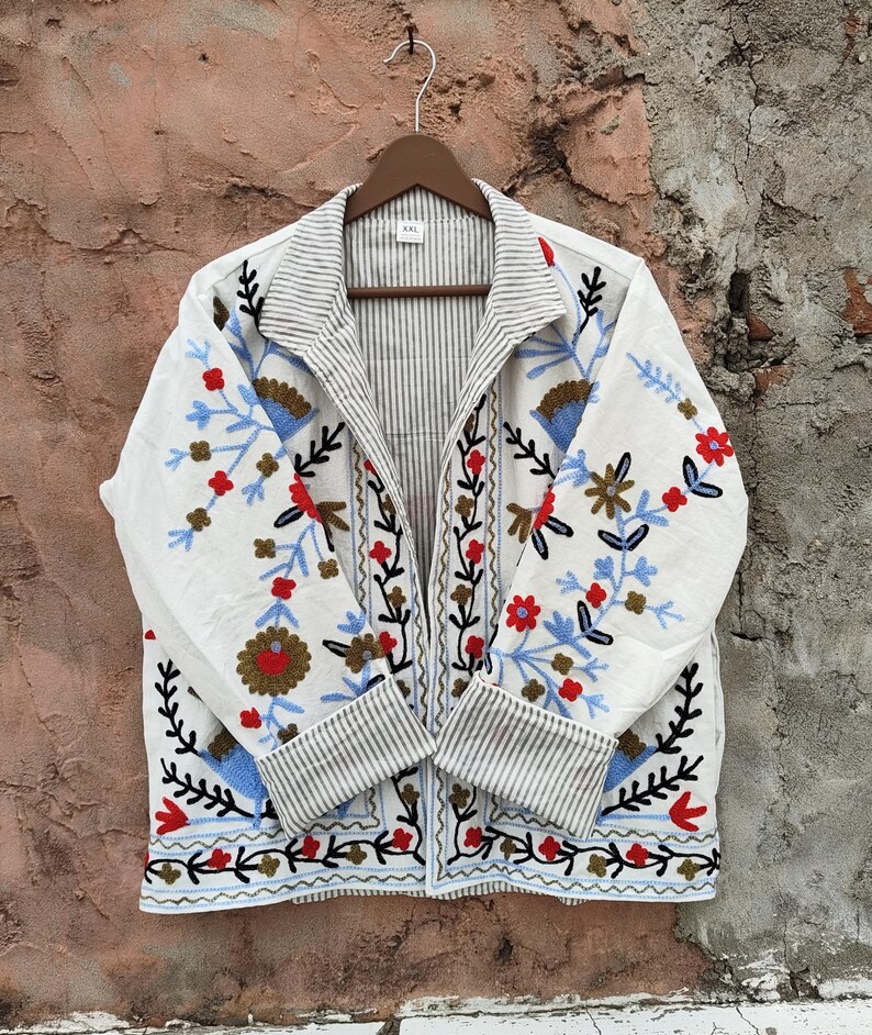 K&ouml;nnte beinhalten: Wei&szlig;e Jacke mit floralen Stickereien in Rot, Blau und Braun. Die Jacke hat ein gestreiftes Futter und Manschetten und h&auml;ngt an einem Holzb&uuml;gel. Die Stickerei zeigt eine Vielzahl von Blumen und Bl&auml;ttern.