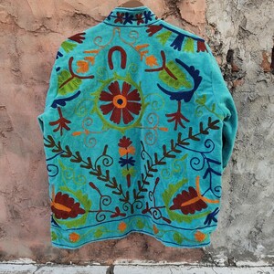 May include: A turquoise jacket with intricate floral embroidery in shades of red, orange, green, and brown. The jacket features a high collar and a detailed design on the back, showcasing a unique, handcrafted style.
