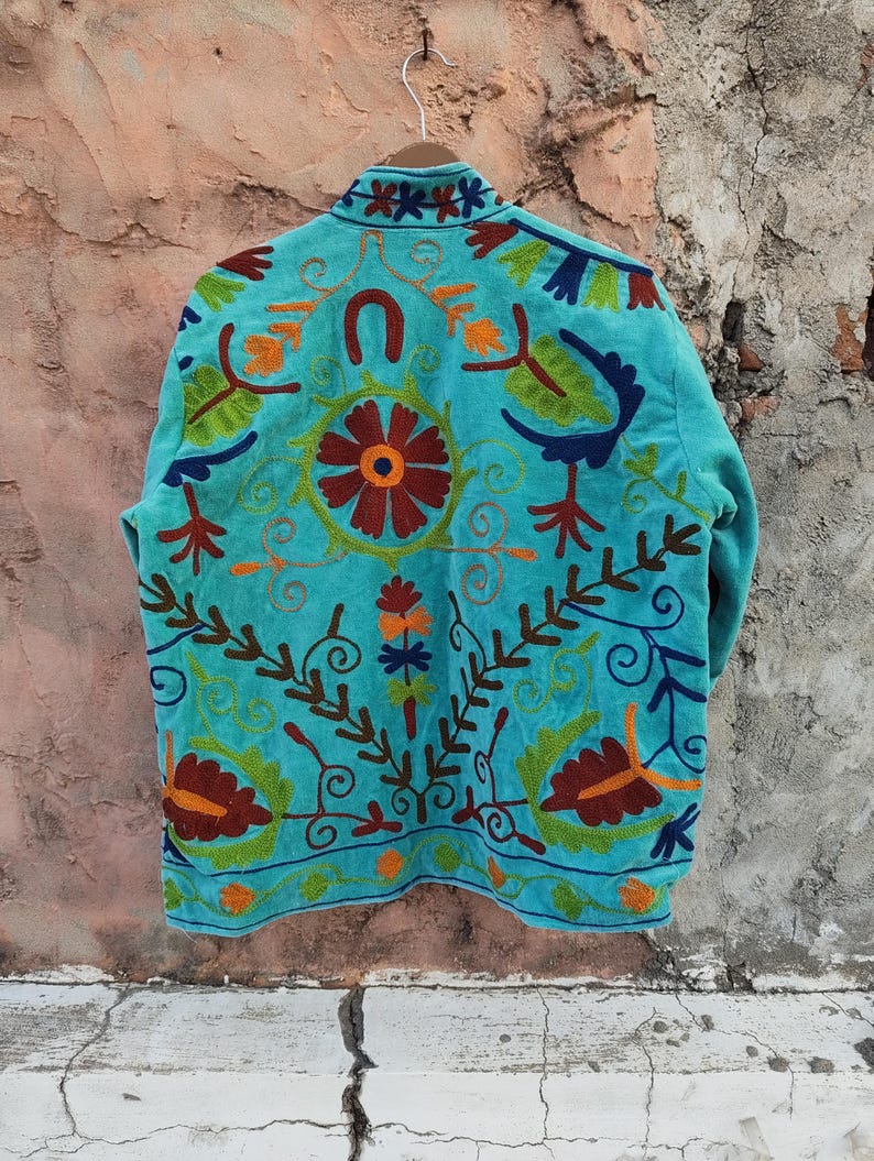 May include: A turquoise jacket with intricate floral embroidery in shades of red, orange, green, and brown. The jacket features a high collar and a detailed design on the back, showcasing a unique, handcrafted style.