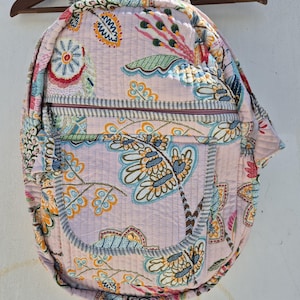 Puede incluir: Una mochila acolchada rosa con estampado floral. La mochila tiene un bolsillo grande en la parte delantera y cierre de cremallera.