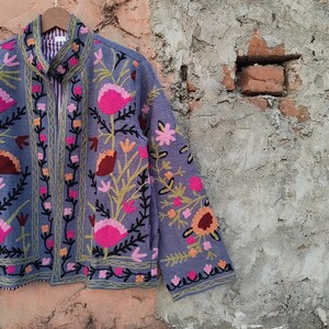 May include: A periwinkle blue jacket with intricate floral embroidery in shades of pink, orange, and green. The jacket features a zip-up front and a striped lining. The floral design is detailed with black outlines, creating a striking contrast against the blue fabric.