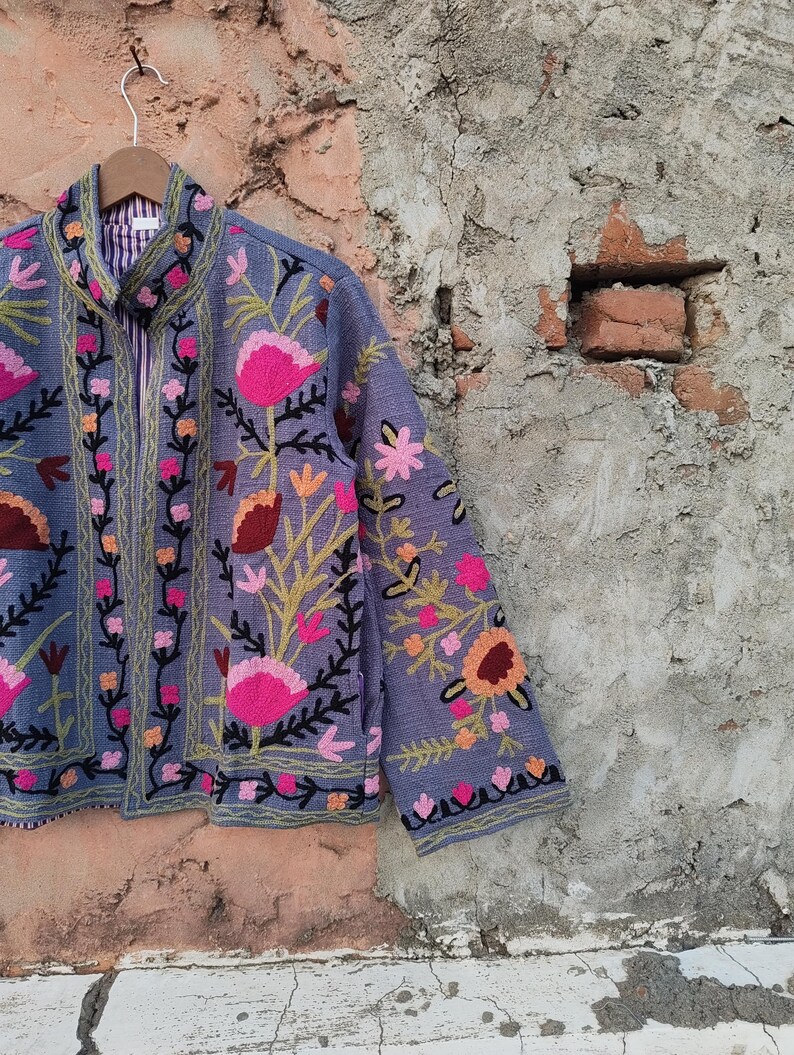 May include: A periwinkle blue jacket with intricate floral embroidery in shades of pink, orange, and green. The jacket features a zip-up front and a striped lining. The floral design is detailed with black outlines, creating a striking contrast against the blue fabric.