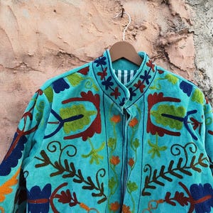 May include: A turquoise jacket with intricate floral embroidery in shades of red, green, orange, and blue. The jacket is hanging on a wooden hanger against a textured background. The embroidery features a detailed, colorful design.