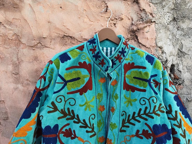 May include: A turquoise jacket with intricate floral embroidery in shades of red, green, orange, and blue. The jacket is hanging on a wooden hanger against a textured background. The embroidery features a detailed, colorful design.