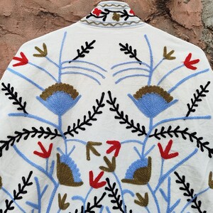 K&ouml;nnte beinhalten: Weisse Jacke mit aufwendiger Blumenstickerei in Blau, Rot, Schwarz und Braun. Das Design zeigt stilisierte Blumen- und Bl&auml;ttermuster, die eine symmetrische Anordnung bilden. Der Kragen der Jacke ist ebenfalls mit Stickereien verziert.