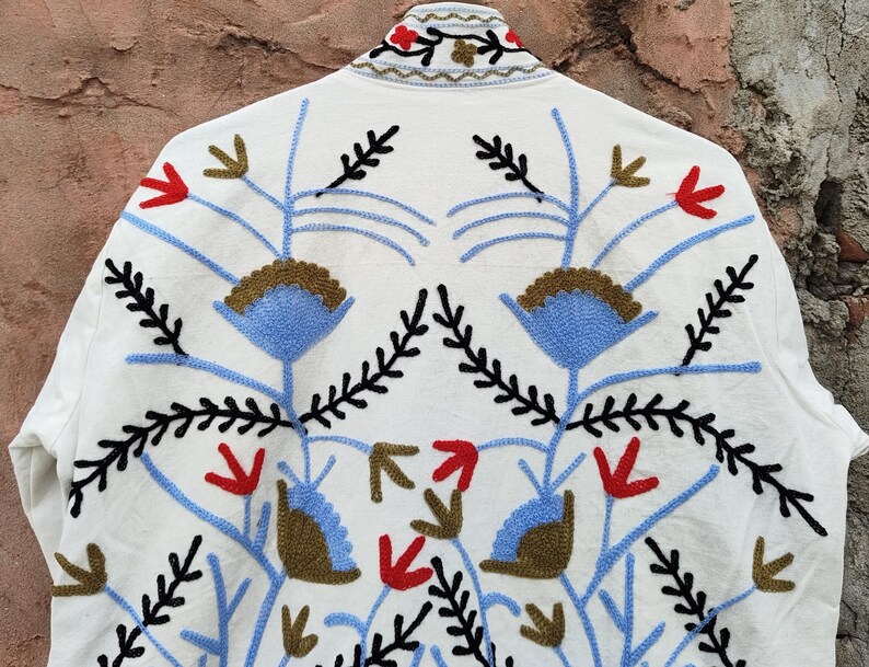 K&ouml;nnte beinhalten: Weisse Jacke mit aufwendiger Blumenstickerei in Blau, Rot, Schwarz und Braun. Das Design zeigt stilisierte Blumen- und Bl&auml;ttermuster, die eine symmetrische Anordnung bilden. Der Kragen der Jacke ist ebenfalls mit Stickereien verziert.
