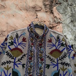 May include: A beige jacket with intricate floral embroidery in blue, red, green, and yellow. The jacket features a mandarin collar and a striped lining. The design includes detailed floral patterns and a vertical border of embroidered elements.