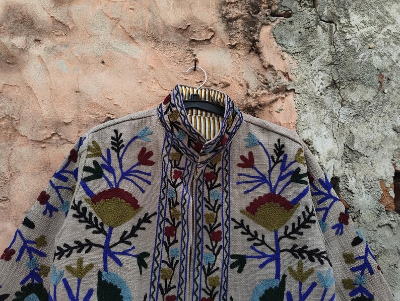 May include: A beige jacket with intricate floral embroidery in blue, red, green, and yellow. The jacket features a mandarin collar and a striped lining. The design includes detailed floral patterns and a vertical border of embroidered elements.