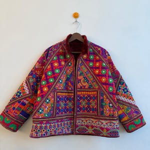 Könnte beinhalten: Eine langärmelige Jacke mit einem lebendigen, mehrfarbigen Patchwork-Design, aufwendigen Stickereien, kreisförmigen Spiegelakzenten und geometrischen Mustern in Magenta, Orange, Grün und Blau, abgerundet durch einen dunkelroten Kragen.