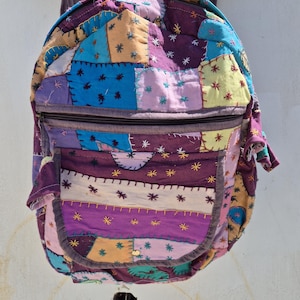 以下が含まれることがあります： 紫、青、黄色、オレンジなど、さまざまな色の生地の正方形を組み合わせたカラフルなパッチワークバックパック。 バックパックには、ジッパー付きのメインコンパートメントとフロントポケットがあります。 デザインには、刺繍された星のディテールと装飾的なステッチが含まれています。