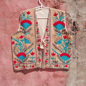 Puede incluir: Un chaleco beige sin mangas con un diseño de bordado floral. El bordado presenta flores rojas, azules y verdes sobre un fondo beige.