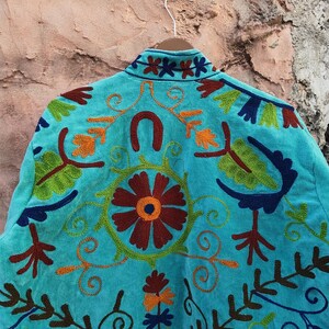 May include: A turquoise jacket with intricate embroidery. The back of the jacket features a large floral design in shades of red, orange, and green, surrounded by swirling patterns and other floral and leaf motifs. The collar is also embroidered.