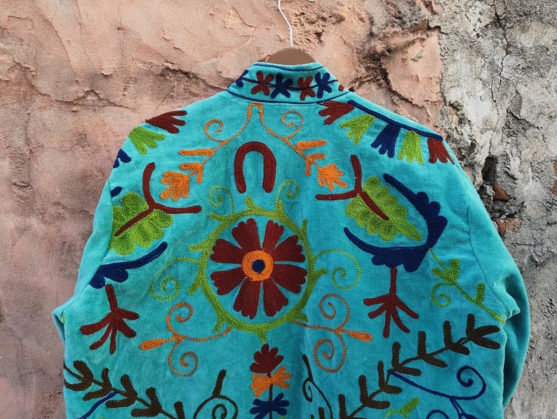 May include: A turquoise jacket with intricate embroidery. The back of the jacket features a large floral design in shades of red, orange, and green, surrounded by swirling patterns and other floral and leaf motifs. The collar is also embroidered.