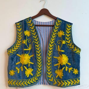 Puede incluir: Un chaleco de terciopelo azul con bordados florales de color amarillo brillante en los paneles frontales y los bordes. La prenda está forrada con una tela a rayas azul claro y blanca y se muestra en una percha de madera marrón.