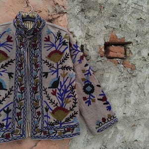 May include: A beige jacket with intricate floral embroidery in blue, red, and green. The jacket has a high collar and a zipper closure. The sleeves and front panels are adorned with detailed patterns, creating a bohemian aesthetic.