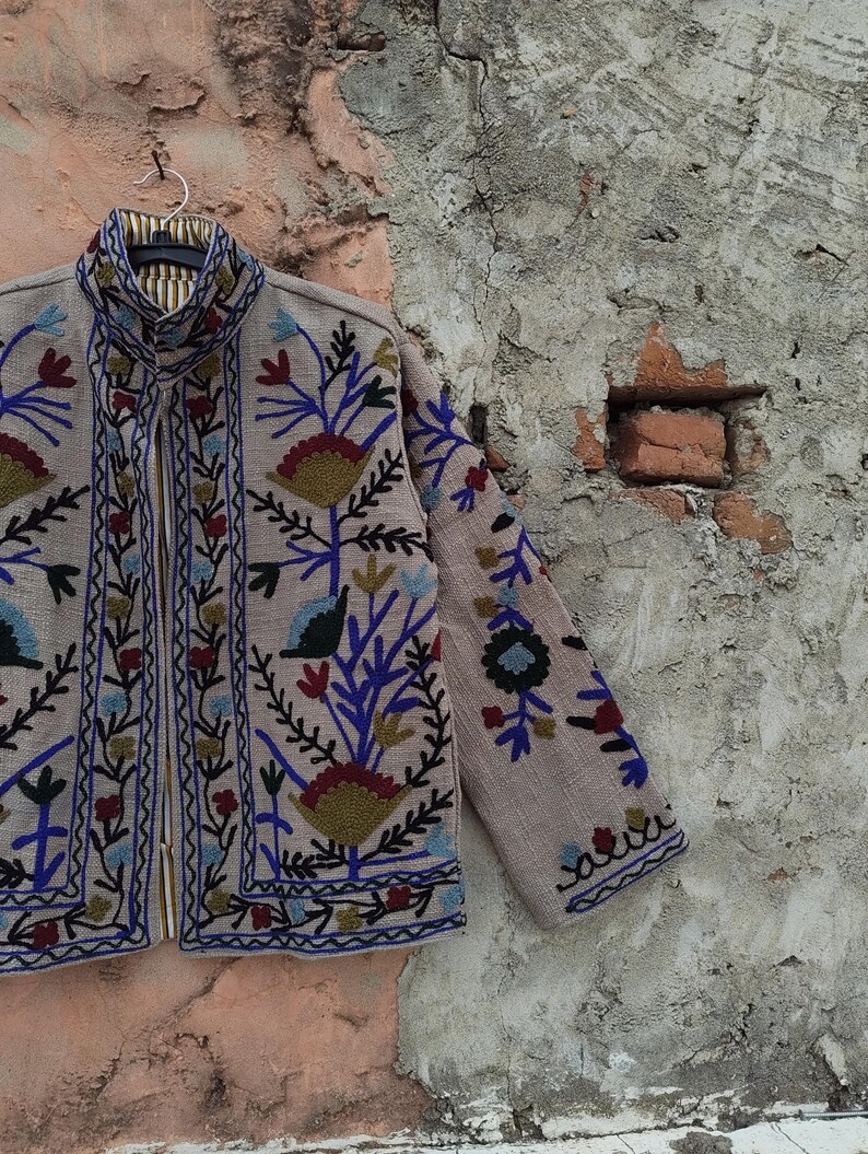 May include: A beige jacket with intricate floral embroidery in blue, red, and green. The jacket has a high collar and a zipper closure. The sleeves and front panels are adorned with detailed patterns, creating a bohemian aesthetic.