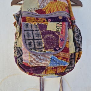 以下が含まれることがあります： パッチワークで作られたバックパック。紫、青、黄、赤の色合いの様々な生地が使われています。バックパックには大きなメインコンパートメントと小さなフロントポケットがあります。ストラップはバックパックと同じ生地で作られています。