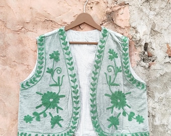 May include: A light green vest with intricate green floral embroidery and leaf patterns. The vest has a striped white and gray lining and is displayed on a wooden hanger. The embroidery details and the vest's design suggest a bohemian or vintage style.
