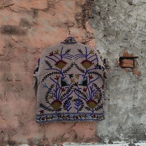 May include: A beige jacket with intricate floral embroidery in red, blue, and green. The jacket is hanging on a metal hanger against a textured, weathered wall. The embroidery features stylized flowers and leaves.