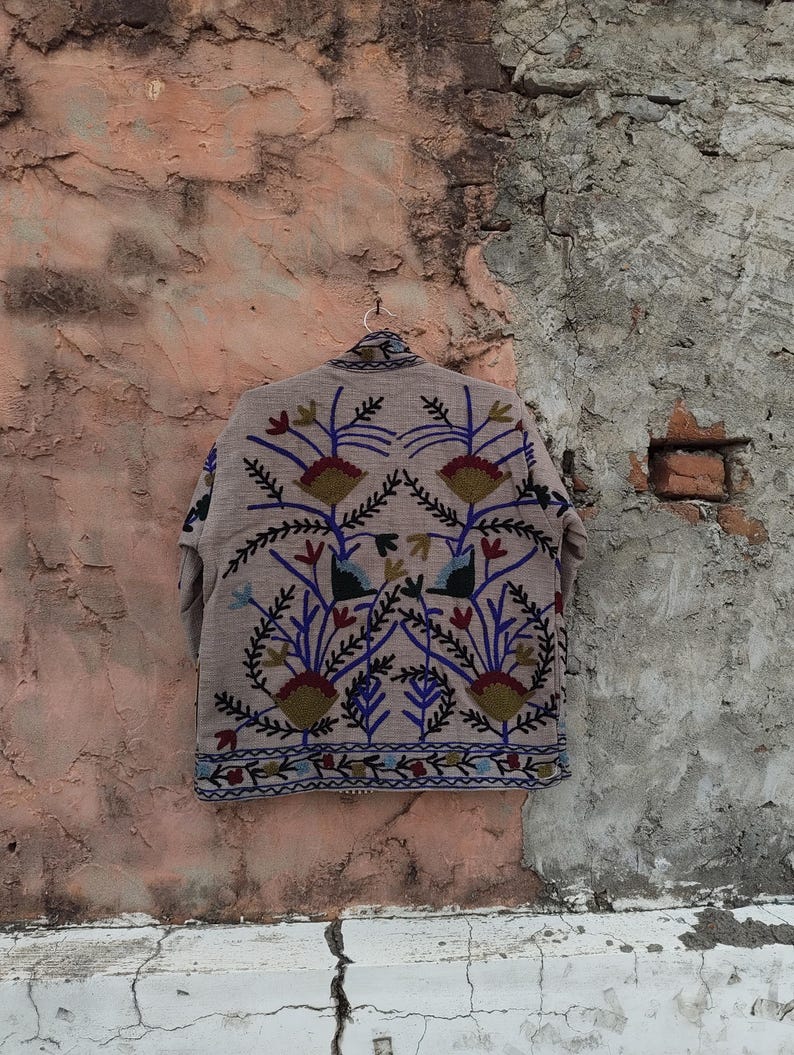 May include: A beige jacket with intricate floral embroidery in red, blue, and green. The jacket is hanging on a metal hanger against a textured, weathered wall. The embroidery features stylized flowers and leaves.