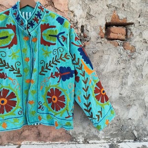 May include: A turquoise jacket with intricate floral embroidery in shades of orange, brown, green, and blue. The jacket is hanging on a wooden hanger against a textured wall. The embroidery details are visible on the collar and sleeves.