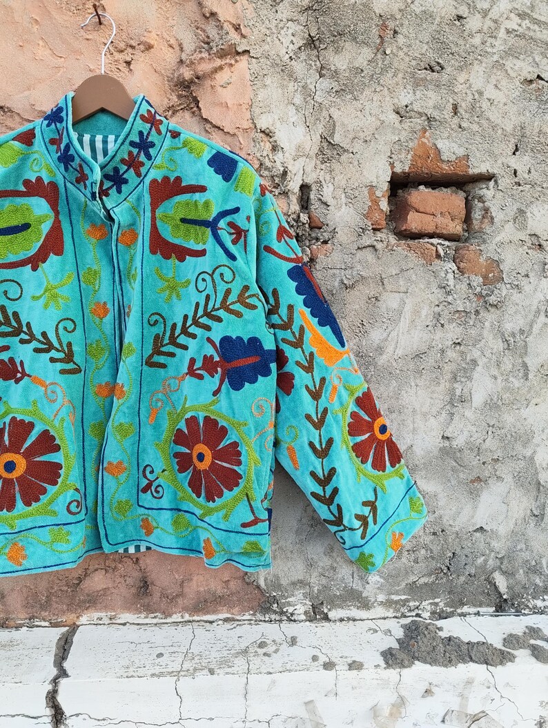 May include: A turquoise jacket with intricate floral embroidery in shades of orange, brown, green, and blue. The jacket is hanging on a wooden hanger against a textured wall. The embroidery details are visible on the collar and sleeves.