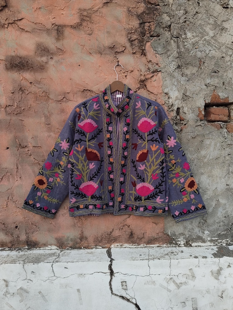 May include: A periwinkle-colored jacket with intricate floral embroidery in pink, orange, and green. The jacket features a stand-up collar and a full-length zipper. The floral design is symmetrical, with detailed stitching.