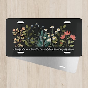 May include: Black license plate featuring a floral design with pink, white, and blue wildflowers. The text "consider how the wildflowers grow" is written in white script across the bottom. The plate has four mounting holes.
