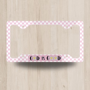 May include: A license plate frame with a pink and white checkered pattern. The frame has the words "GOD IS GOOD" in a pink rectangle with daisy accents. The frame is designed to fit a standard U.S. license plate.