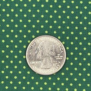 May include: A dark green fabric featuring a repeating pattern of small, light green polka dots. A silver United States quarter coin is placed on top of the fabric to provide a sense of scale for the size of the printed dot pattern.