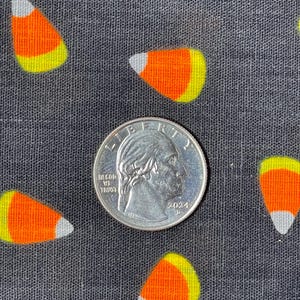May include: Close-up of a quarter coin next to several candy corn designs on a dark gray fabric. The candy corn pieces are orange, yellow, and white. The quarter shows the profile of George Washington and the year 2024.