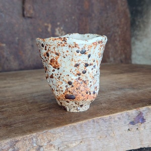 Puede incluir: Una taza pequeña de cerámica blanca con un esmalte marrón moteado. La taza tiene una forma redondeada y un borde ligeramente acampanado.