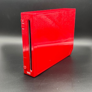 Könnte beinhalten: Eine rote Nintendo Wii-Konsole mit strukturierter Oberfläche. Die Konsole verfügt über einen Schlitz für Spielscheiben und Tasten für Ein- und Ausschalten sowie zum Zurücksetzen.