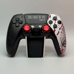 May include: A black and white PlayStation 5 DualSense controller with red buttons. The controller has a custom design with a white background and black and red splatters. The text "THE LAST OF US PART II" is printed on the top of the controller.