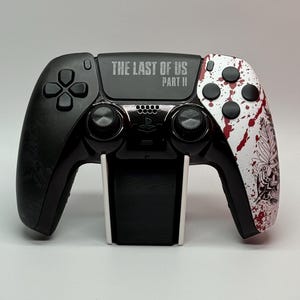 May include: A black and white PlayStation 5 controller with a red and white splatter paint design. The controller has the text "THE LAST OF US PART II" printed on the top.