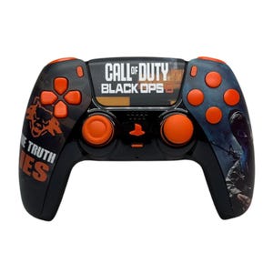 Könnte beinhalten: Ein schwarzer PlayStation 5-Controller mit orangenen Knöpfen und einem Call of Duty: Black Ops Cold War-Design. Der Controller verfügt über ein schwarz-oranges Tarnmuster mit dem Spiellogo und dem Schriftzug "Embrace the Truth" auf der linken Seite.