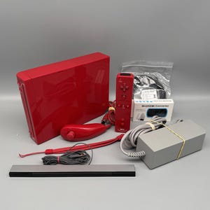 May include: A red Nintendo Wii console with matching red remote controllers, a sensor bar, and power supply. Includes a Wii2HDMI converter and various cables. The console and accessories are arranged on a gray surface.