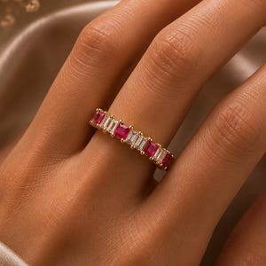 以下が含まれることがあります： 長方形にカットされた赤いルビーと透明なバゲットカットの白い石が交互に並ぶゴールドのバンドリング。金属製のバンドは光沢のある仕上げで、共有爪留めによって宝石がしっかりと固定されています。